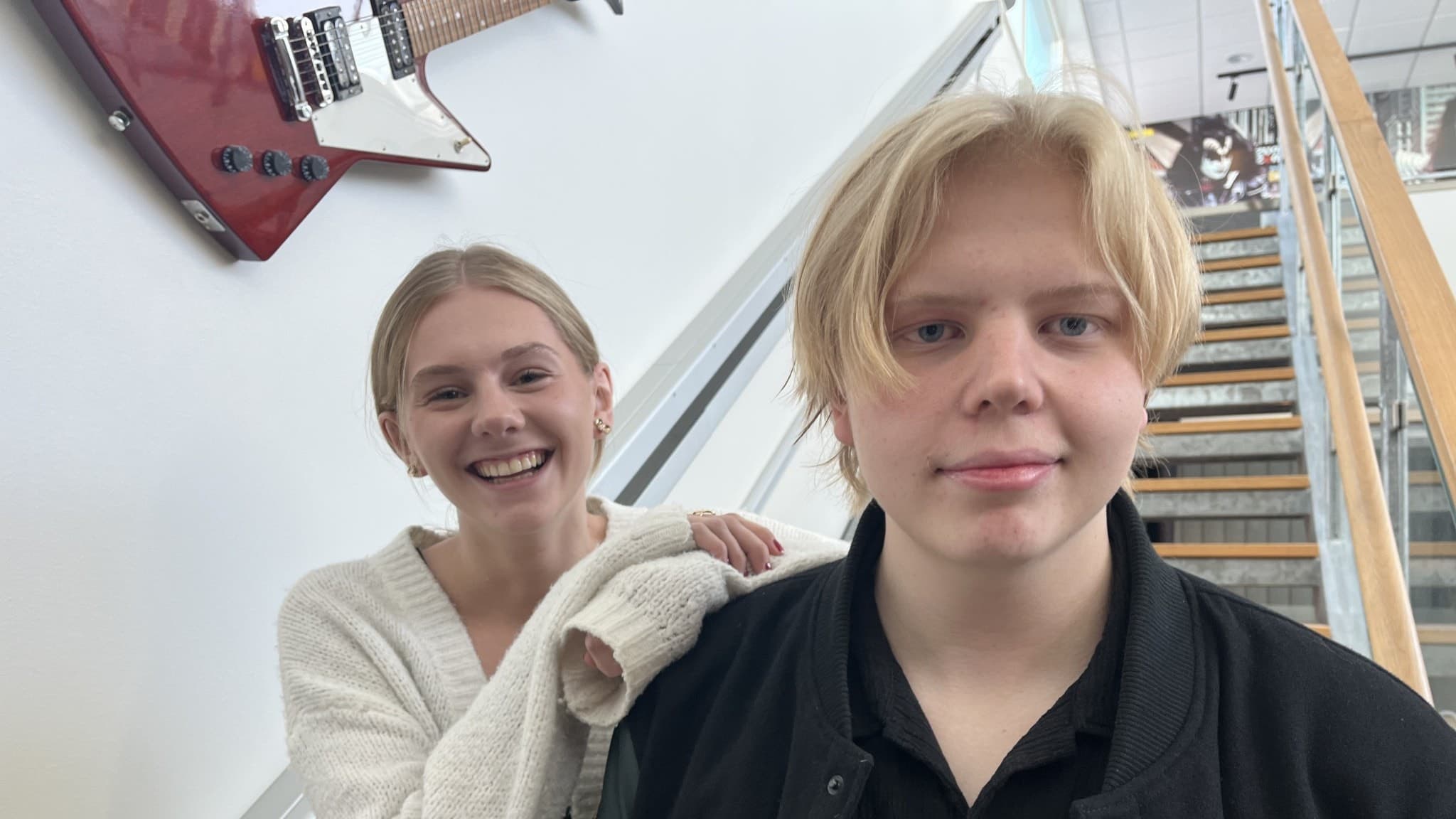 Två personer står framför en trappa inomhus. I bakgrunden syns en vägg med en röd gitarr som dekoration. Den ena personen bär en svart jacka med broderat namn.