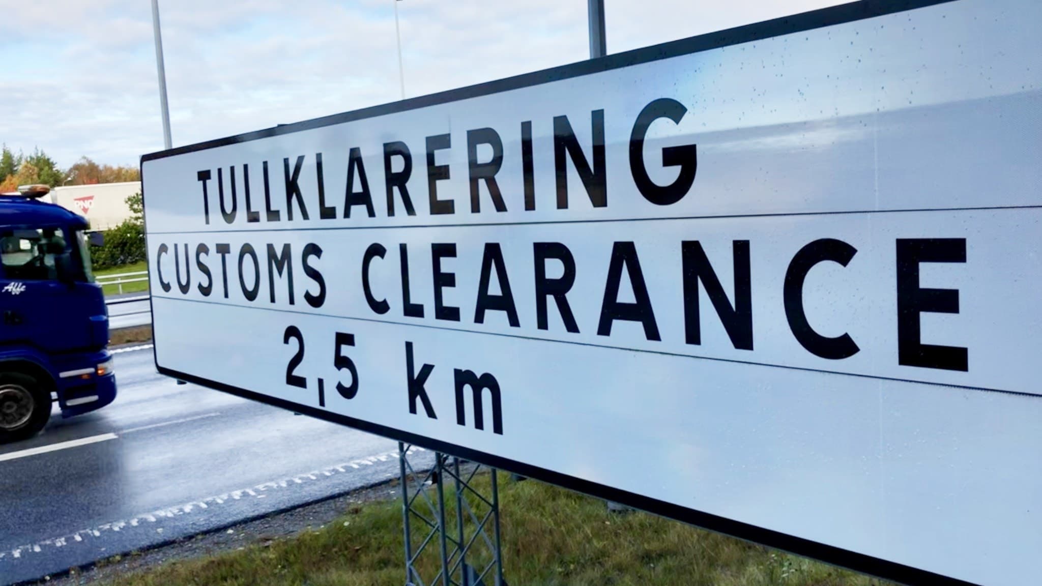Skylt vid Svinesund visar vägen till tullklarering 2,5 km framåt. I bakgrunden syns en lastbil på en väg omgiven av grönskande landskap.