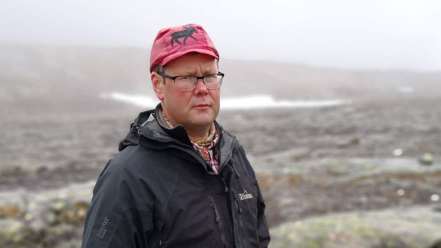 Anders-Erling Fjällås, ordförande i Semisjaur-Njarg sameby, står utomhus i fjällmiljö med dimmig bakgrund och klädd i svart jacka och röd mössa med motiv.