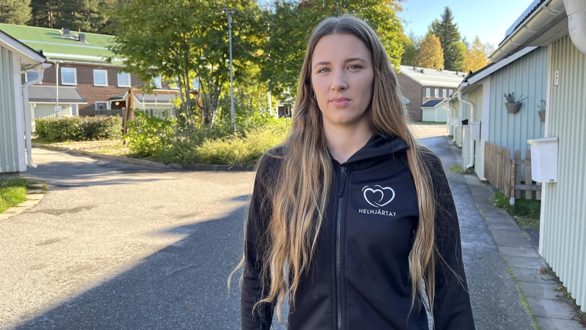 Kvinna med långt blont hår står utomhus på en lugn gata med trähus och grönska i bakgrunden. Hon bär en svart hoodie med ett tryck av ett hjärta på bröstet.