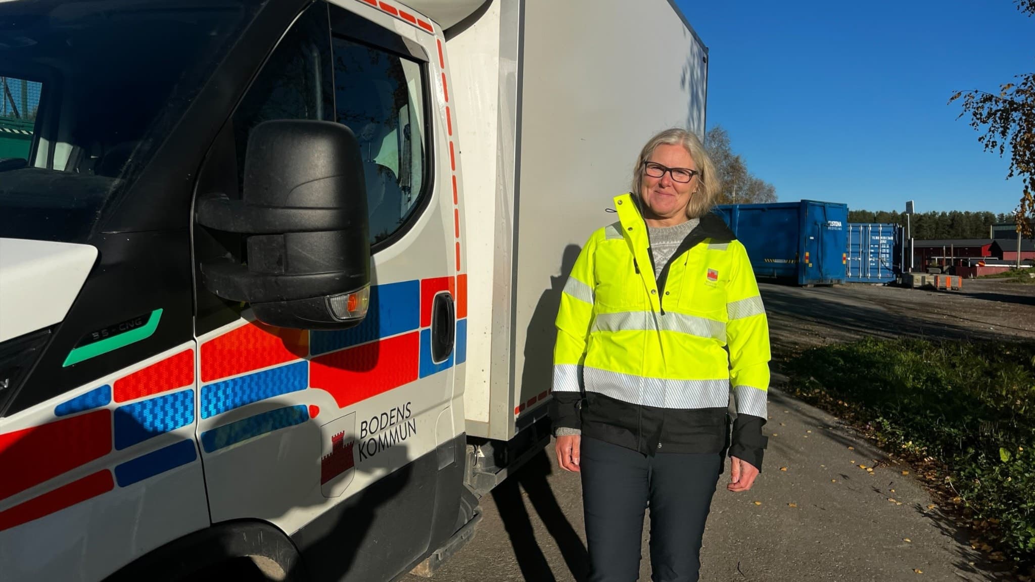 Ulrika Jonsson, avfallshandläggare på Bodens kommun, står framför en lastbil i arbetskläder. Solen skiner och det syns containrar i bakgrunden på en arbetsplats.