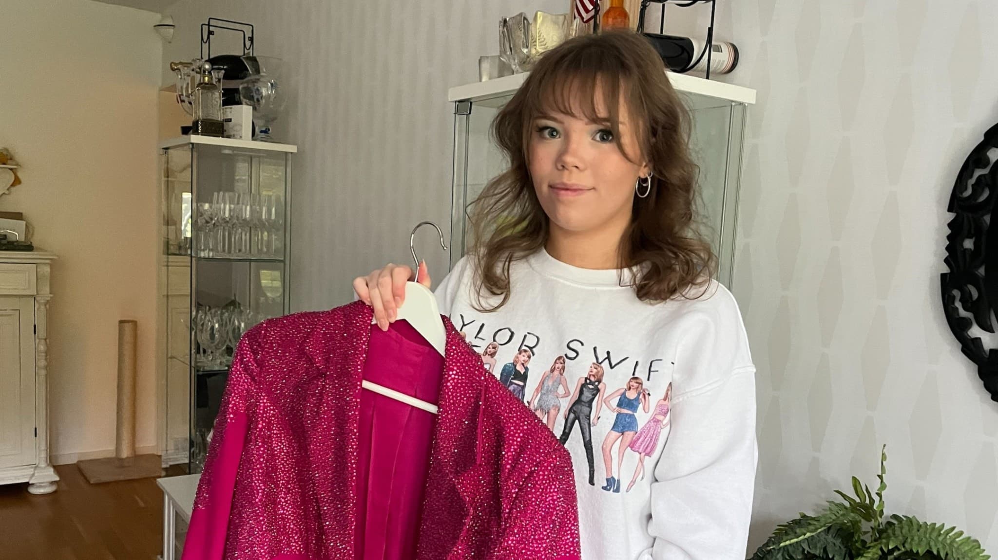 Elin Johansson håller upp en glittrande rosa jacka i ett hem med vit inredning. Hon bär en vit tröja med tryck som hyllar Taylor Swift, och visar sin entusiasm inför albumsläppet.