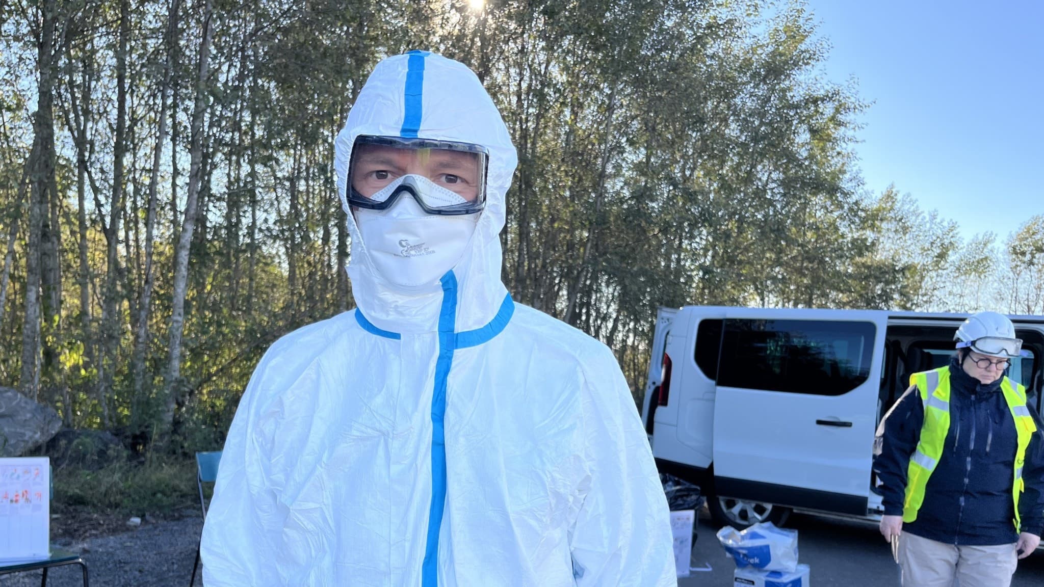 Person klädd i vit skyddsdräkt med blå detaljer står utomhus nära en vit transportbil. En annan individ i gul väst och skyddsdräkt syns i bakgrunden. Miljön är omgiven av träd.