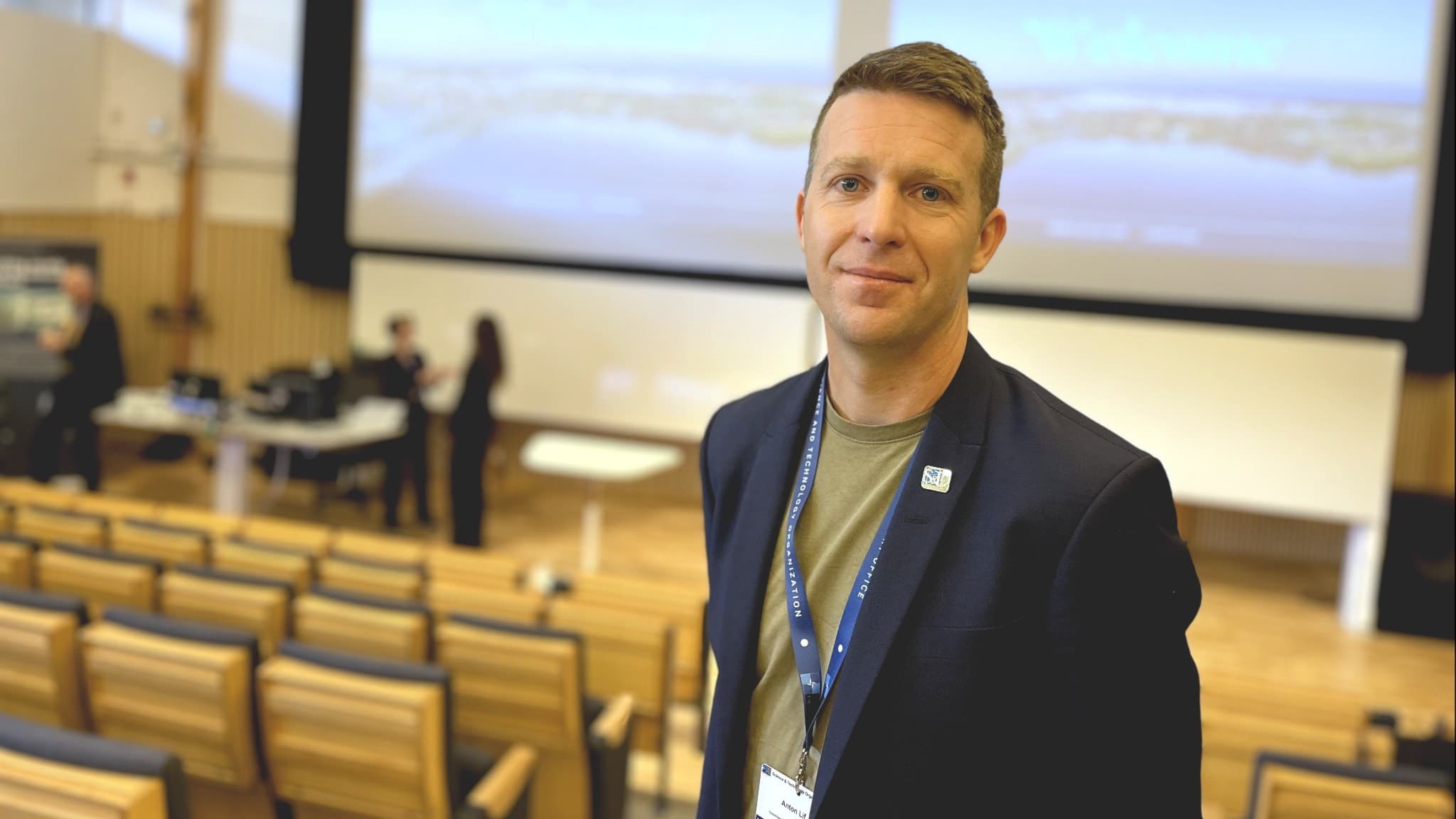Anton Lif står i en föreläsningssal med tomma stolar i förgrunden och en stor presentationsduk i bakgrunden. Personen har en mörk kavaj.
