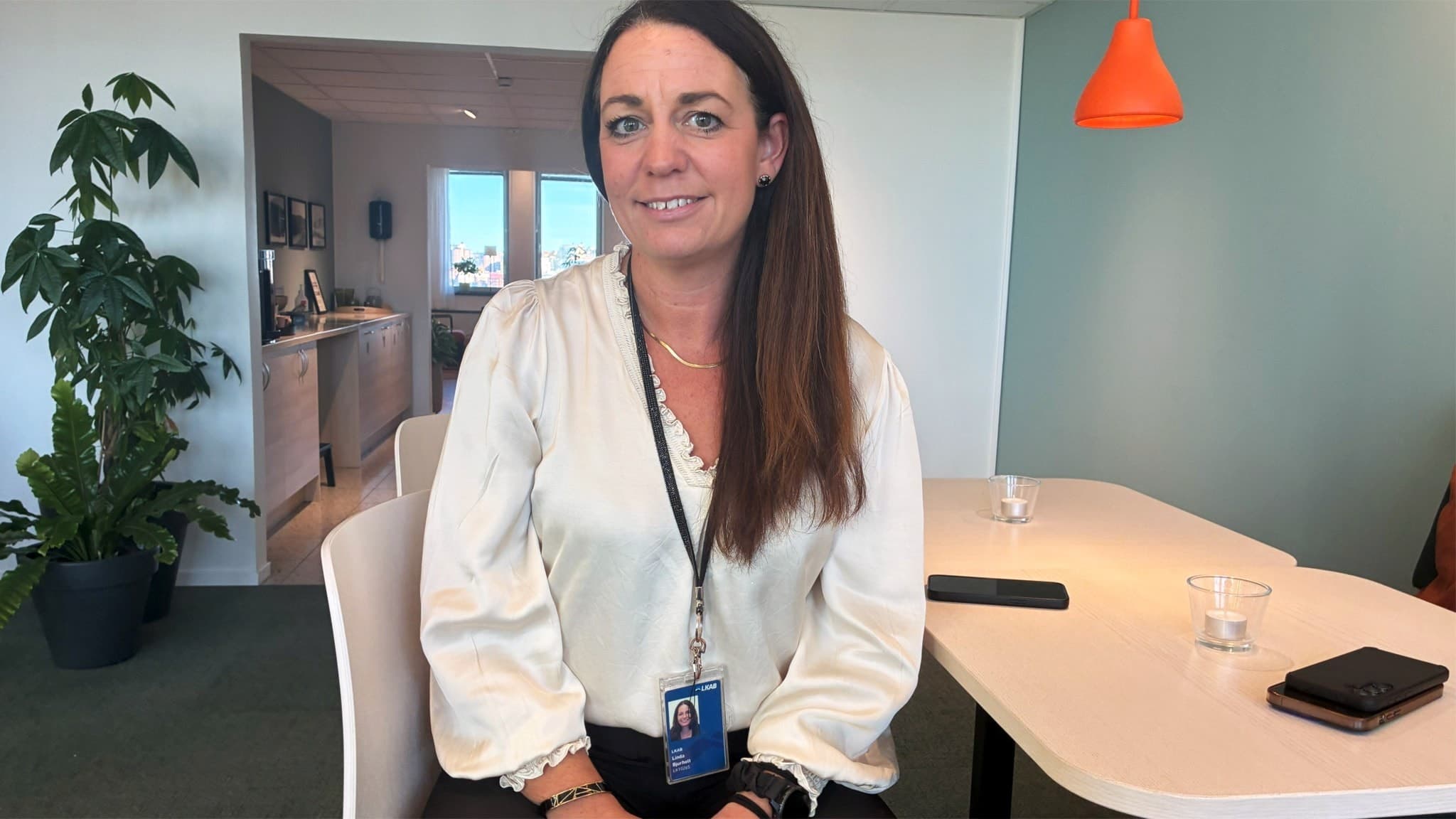 Linda Bjurholt, logistikchef på LKAB, sitter vid ett bord i en modern kontorsmiljö med ljusa väggar och en orange lampa i bakgrunden. På bordet finns glas och en mobiltelefon.
