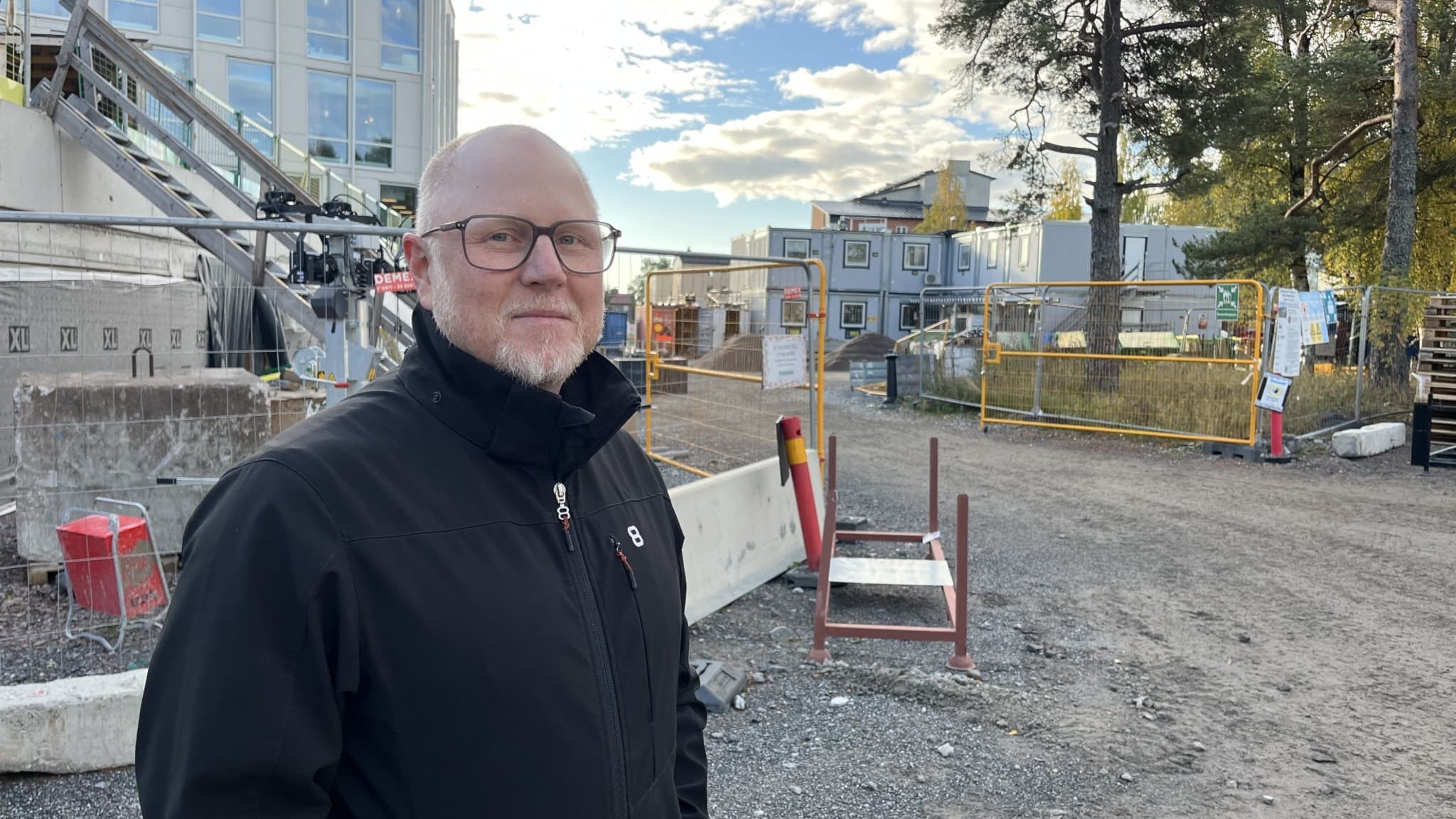 En byggarbetsplats i Luleå med grusväg, byggmaterial och maskiner. En man i svart jacka står nära arbetsområdet. Flera byggnader och en blå himmel med moln syns i bakgrunden.