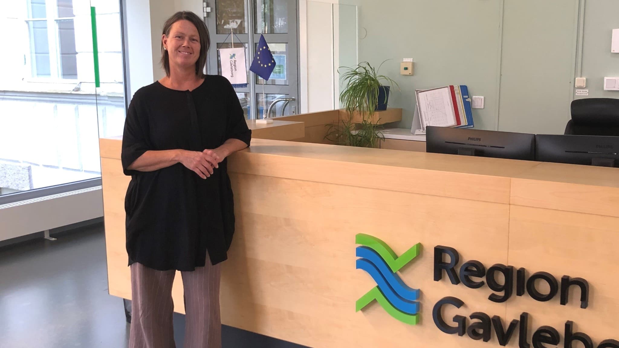 Jennie Forsblom från Kristdemokraterna står vid en receptionsdisk med Region Gävleborgs logotyp i en kontorsmiljö. Bakom henne syns glaspartier och en växt på disken.