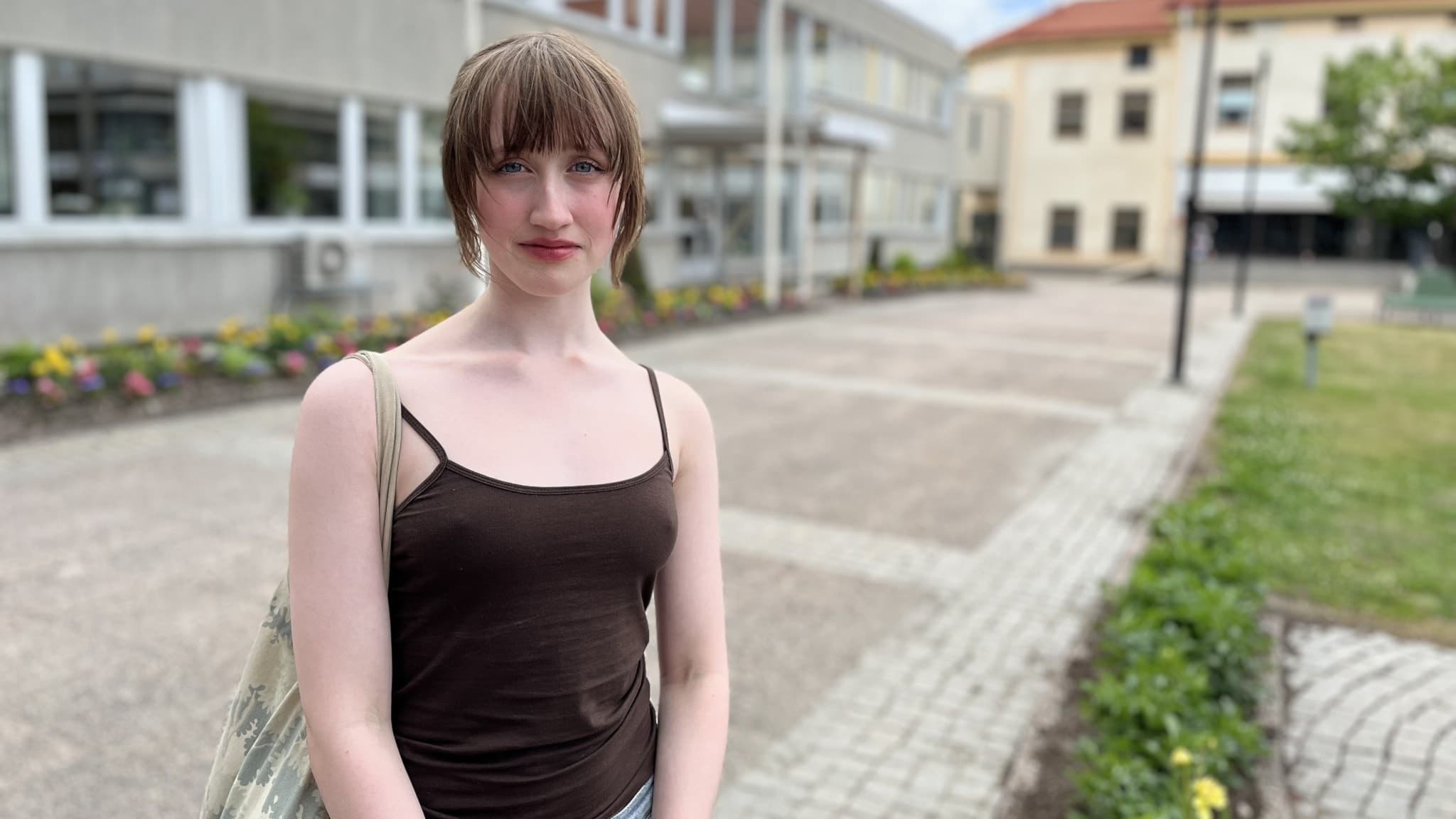 17-åriga Meja Francis Bjärne står utomhus på en skolgård med grå byggnader och gröna rabatter i bakgrunden, klädd i ett brunt linne och jeans, med en axelväska över ena axeln.