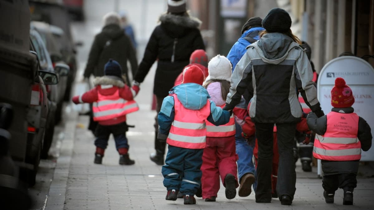 En grupp barn i färgglada reflexvästar går på en trottoar tillsammans med vuxna. Barnen verkar vara från en förskola och leds av pedagoger i vinterkläder.