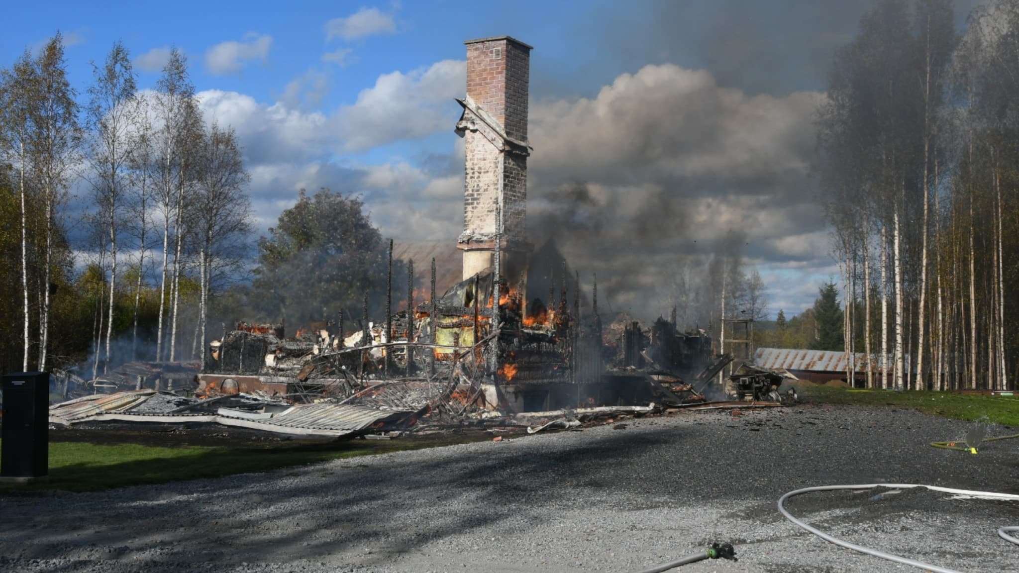 En villa i Färila har totalförstörts i en omfattande brand. Bilden visar rester av byggnaden, med rök och brandhärjade strukturer omgivna av träd under en klar himmel.