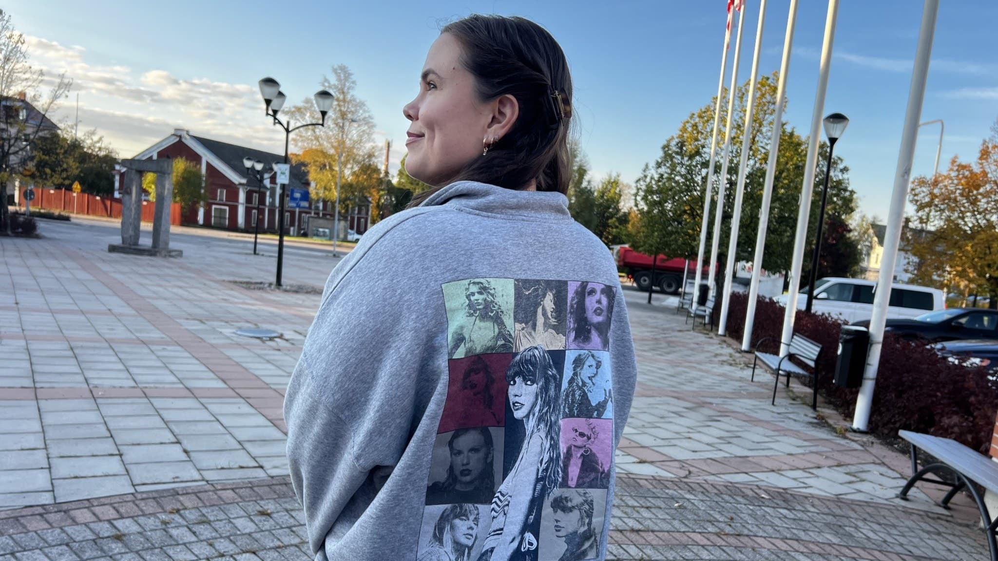 Joline Rudklint från Delsbo står utomhus i en grå tröja med tryck av artisten Taylor Swift på ryggen, omgiven av flaggstänger och en öppen torgmiljö.