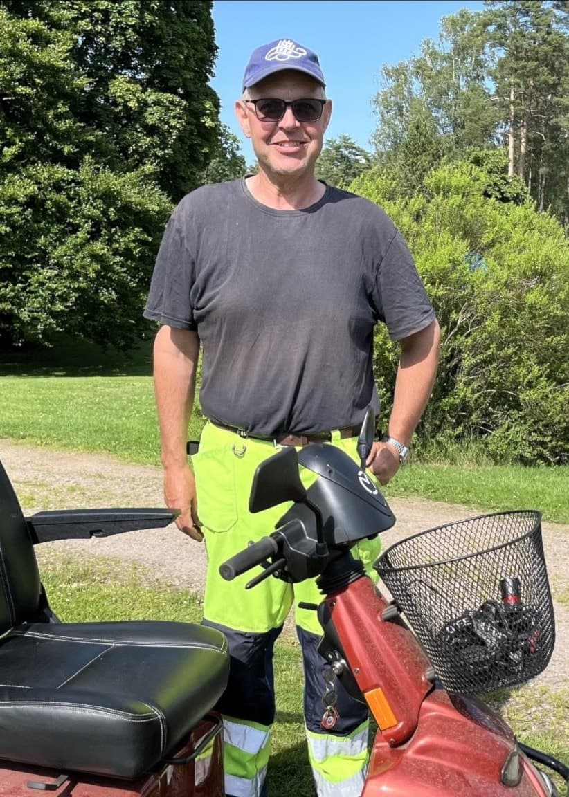 Anders Pettersson står bredvid en röd el-scooter i en grönskande park med buskar och träd i bakgrunden, klädd i arbetskläder och keps.