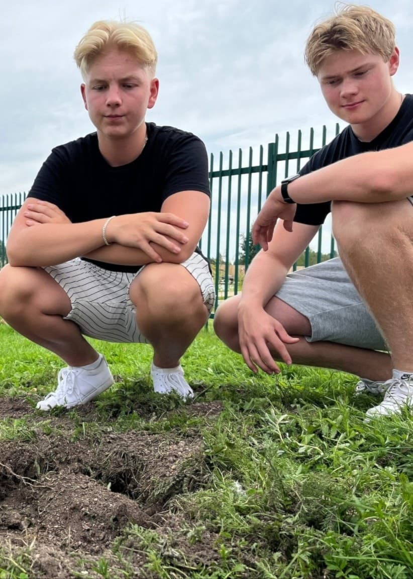 William vid en gräsmatta nära en grop där en rostig sjömina hittats. Sjöminen visas i detalj på vänstra sidan av bilden. Bakgrunden inkluderar staket och vatten.