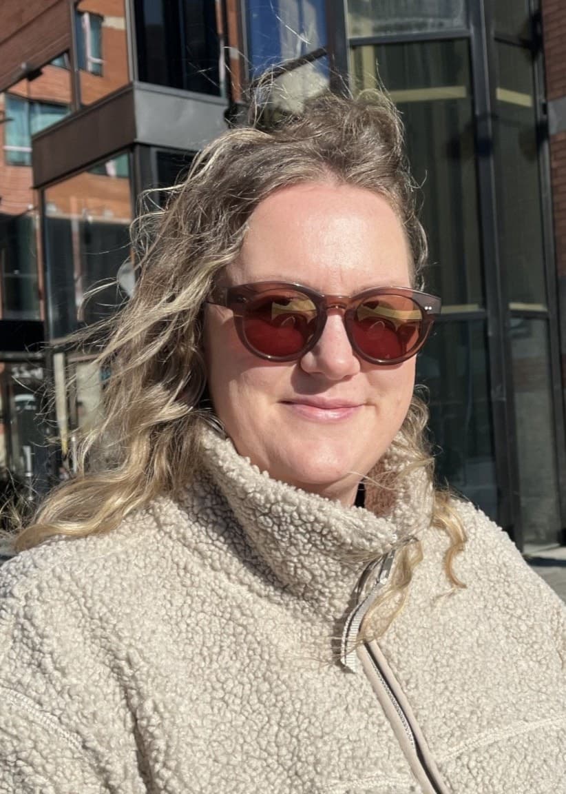 Alexandra Bjur står utomhus i centrala Västerås framför moderna byggnader i glas och tegel. Hon bär en ljus fleecejacka och det är soligt väder.