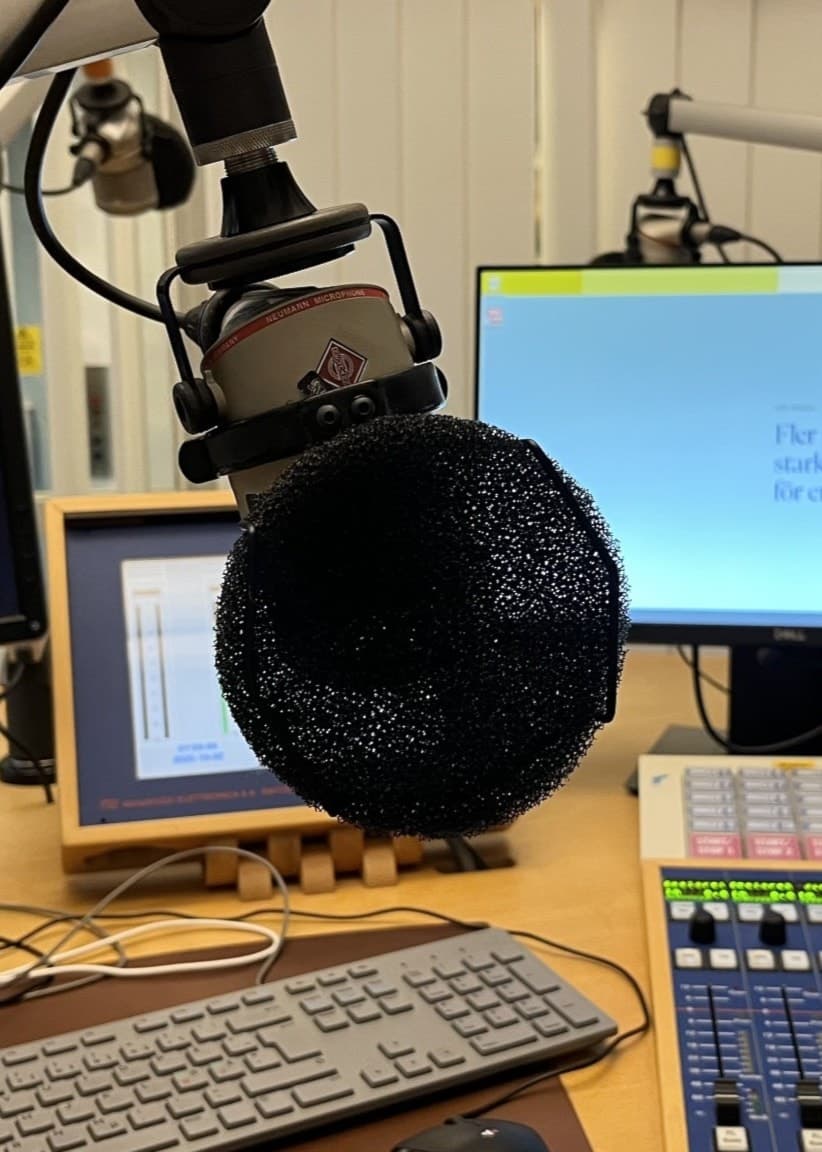 En radiostudio med en mikrofon i förgrunden, flera datorskärmar, ett tangentbord och ljudmixers synliga på ett skrivbord.