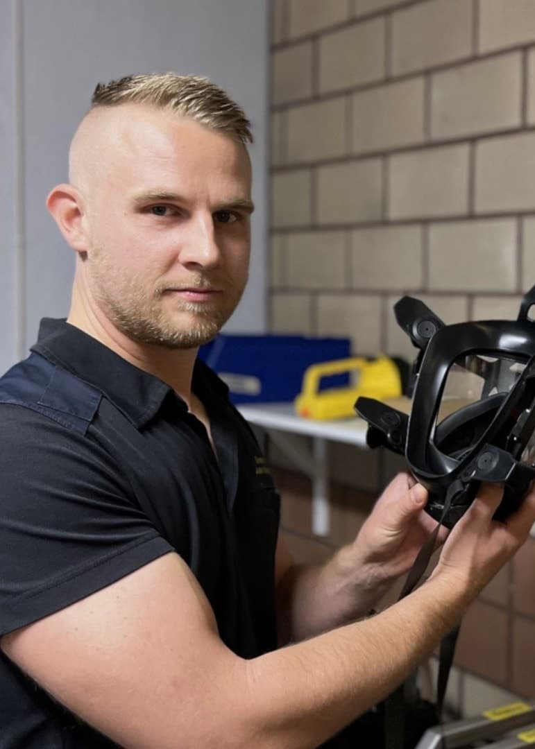 Johan Blomberg, brandman, håller upp en svart andningsmask i en lokal med tegelvägg och informationstavla. Masken används för skydd vid insatser i farliga miljöer.
