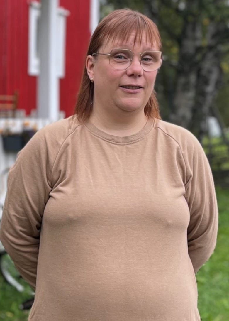 En kvinna med rött hår och beige klänning står framför ett rött hus med vita knutar.