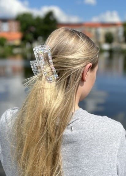 En kvinna med långt blont hår, uppsatt med en hårklämma, står vid en stilla sjö och tittar bort. I bakgrunden syns färgglada byggnader och en blå himmel med moln.