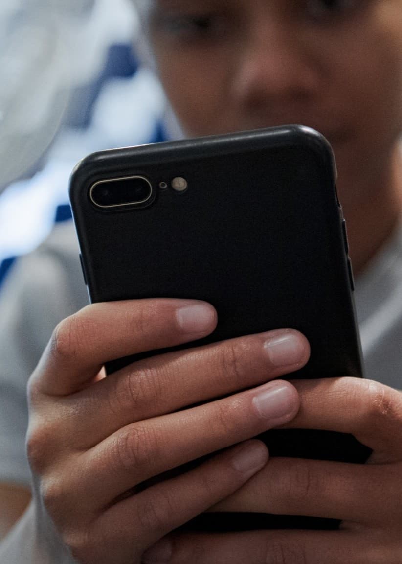 En person håller en mobiltelefon med båda händerna framför ett oskarpt mönstrat bakgrund. Fokus ligger på mobiltelefonen och händerna.