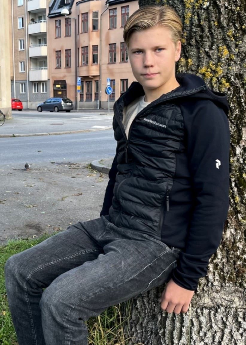 Oskar Leeman lutar sig mot ett träd på en gräsmatta i stadsmiljö med byggnader i bakgrunden. Han bär svart jacka och grå byxor. Bilden visar en lugn scen vid en stadsgata.
