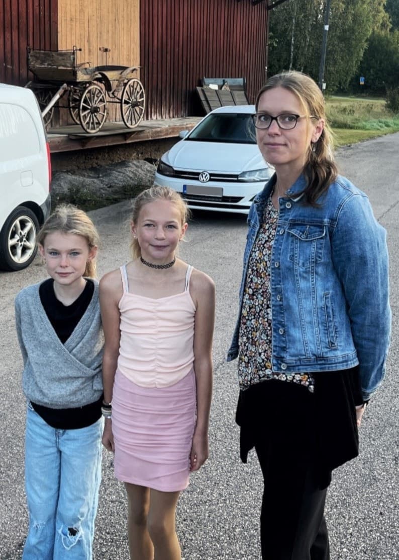 Fyra personer står bredvid en landsväg med en vit skåpbil och en gammal järnvägsvagn i bakgrunden. Landskapet består av grönska och en solnedgång i horisonten.