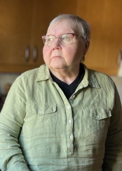 Ulla Svensson står i ett kök med träskåp och en diskho i förgrunden. En vattenkokare syns på bänken bredvid henne, och köksfönstret släpper in dagsljus.