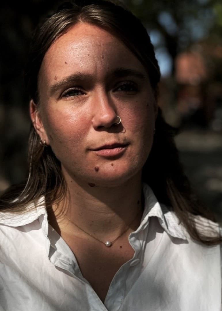Delad bild. Till vänster syns Alba Mogensen, kulturchef på Ystad Allehanda, som står utomhus i en parkmiljö med träd och skuggor i bakgrunden. Hon har på sig en vit skjorta. Till höger syns en person som scrollar på en smartphone. 