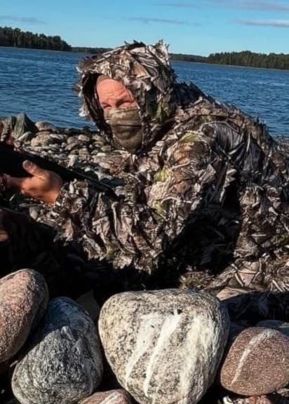 Skyddsjägaren Mattias Landin, klädd i kamouflagedräkt, siktar med gevär vid den Sörmländska kusten. Han är omgiven av stenar och vegetation med havet i bakgrunden.