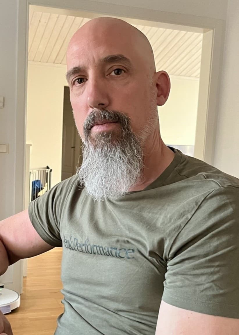 Bild p en man som sitter vid ett köksbord och som har en grön t-shirt på sig. Inget hår men skägg. 