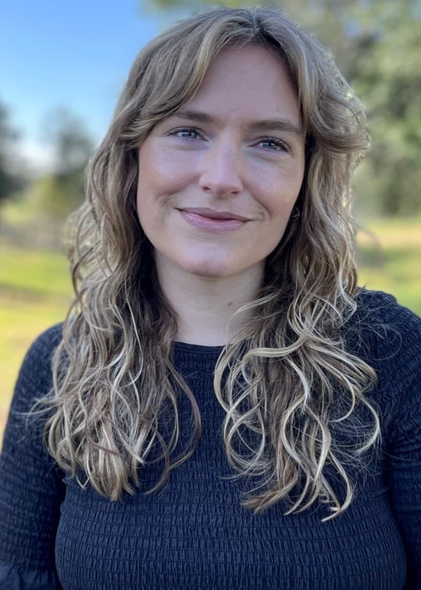 Julia Kvarnberg står utomhus i en grönskande park med träd och blå himmel i bakgrunden. Hon har lockigt blont hår och bär en mörk tröja.