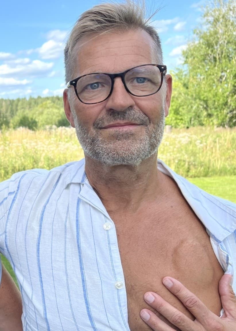 Peter och Nina Eklund står utomhus på en gräsmatta med skog och blå himmel i bakgrunden. Peter visar sitt bröst där en ICD-pacemaker har opererats in.