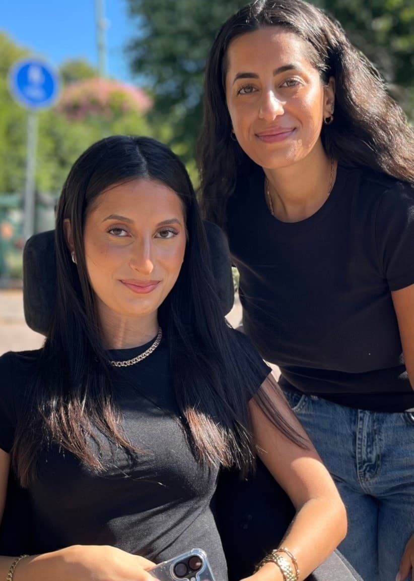 Suzi Bedros, sittande i rullstol, tillsammans med systern Alexandra. De är utomhus en solig dag med träd och en gata i bakgrunden.