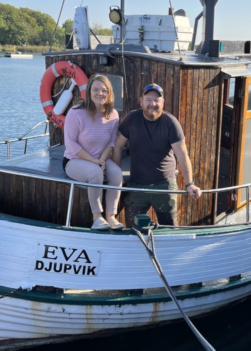 Karoline Andersson och Johan Larsson står på däck av båten Eva, vid kajen i Djupvik. Solen lyser över vattnet och båten, som har en vit skrov med namnet tydligt synligt.