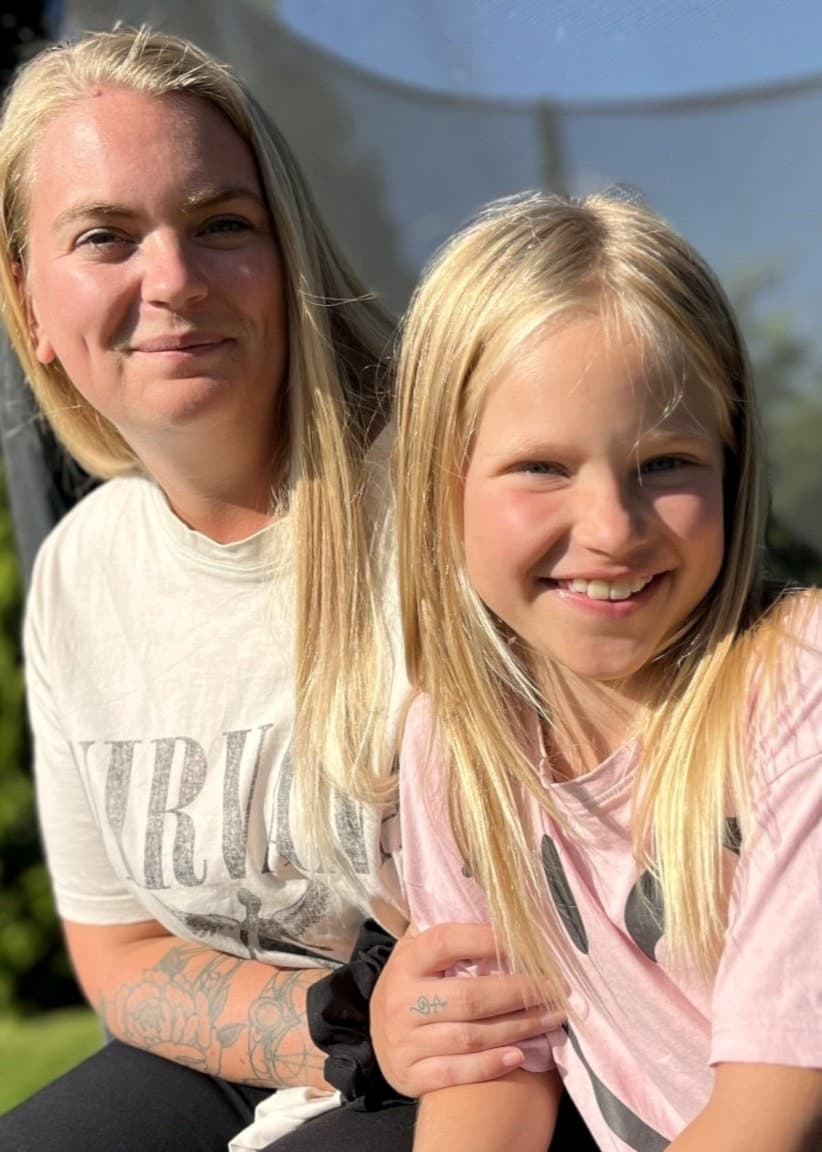 Ronja Luff och hennes dotter Juni sitter tillsammans utomhus på en studsmatta i solskenet. Grönskande buskar och blå himmel syns i bakgrunden. Juni har återhämtat sig efter en sjukdomsperiod.