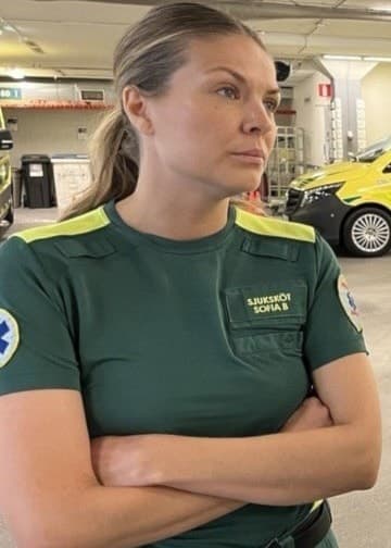 Två ambulanssjukvårdare i gröna uniformer står i en ambulanshall med gula ambulansfordon i bakgrunden och diskuterar arbetsrelaterade frågor.