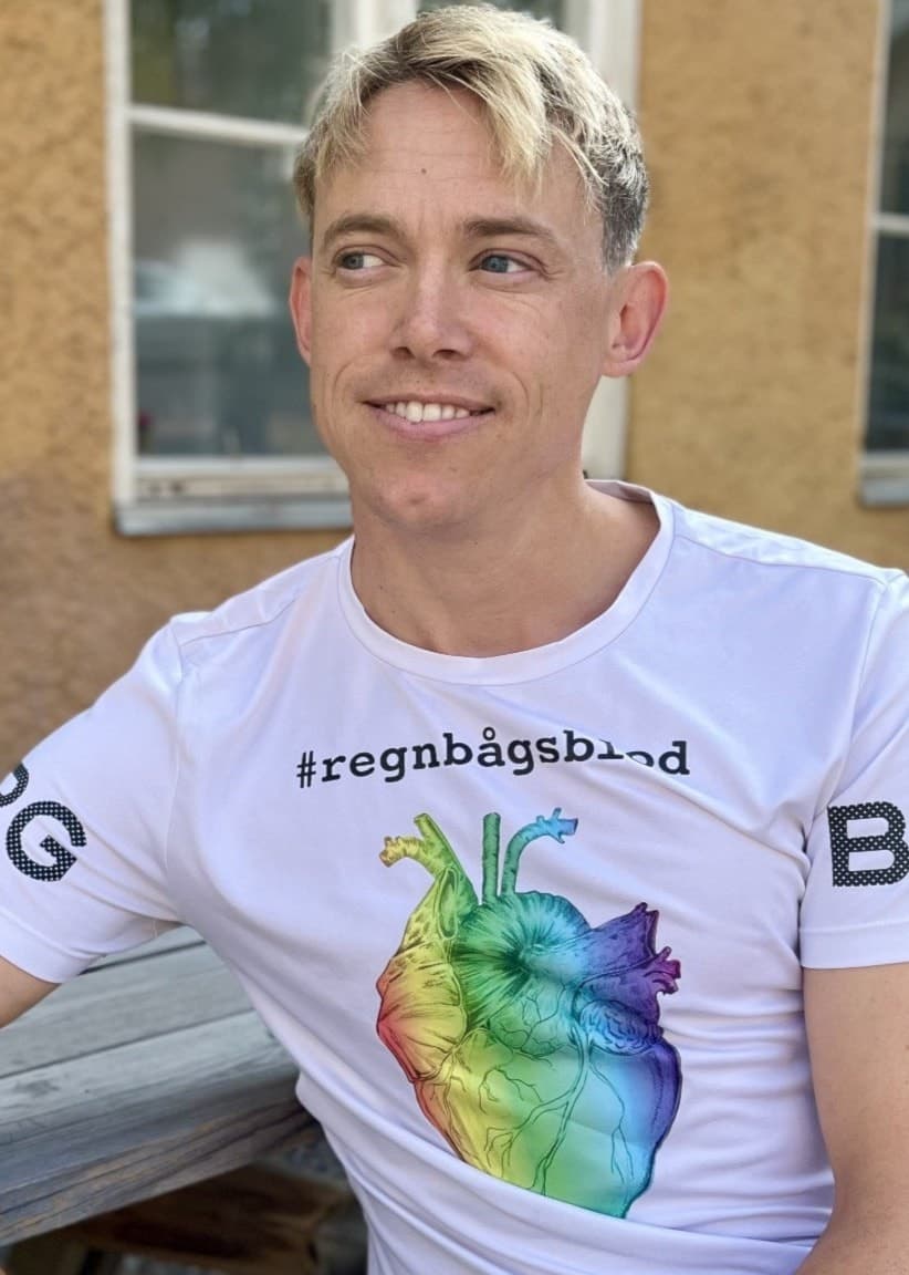 En ung, blond man, sjuksköterskan Tobias Ström, sitter utomhus på en bänk. Han bär en vit T-shirt med en regnbågsfärgad illustration av ett hjärta och texten #regnbågsblod som framhäver hbtq-rättigheter.
