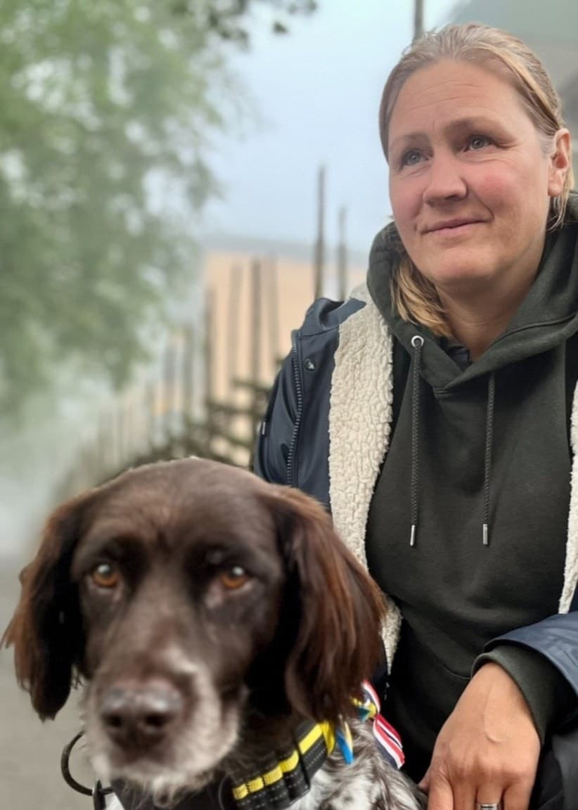 Lotta Nordlöf sitter på en bänk vid en trästaket, klädd i mörka kläder och en jacka med foder. Vid hennes sida sitter en brunvit hund med sele. Bakgrunden visar en dimmig stadsmiljö.