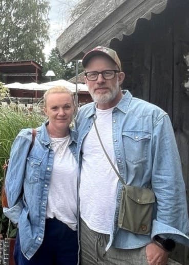Två personer står framför en röd stuga med vita fönster och grön fönsterlucka, omgivna av grusgång, blommor och grönska. Bilden visar en lantlig miljö vid Tovastugan.