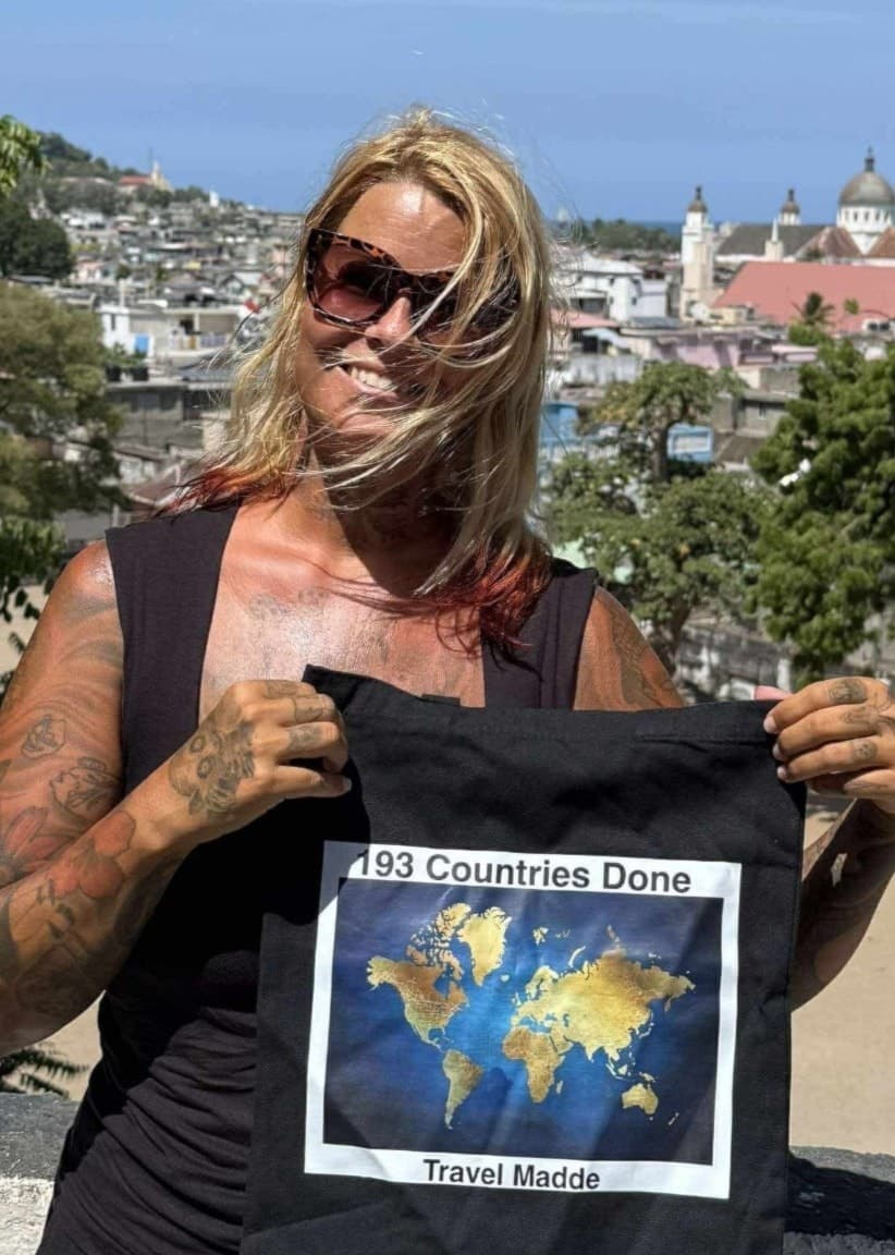 Madde Karlsson står utomhus med en stad i bakgrunden och håller upp en svart t-shirt med en världskarta tryckt på. Soligt väder och en kyrka syns i fjärran.