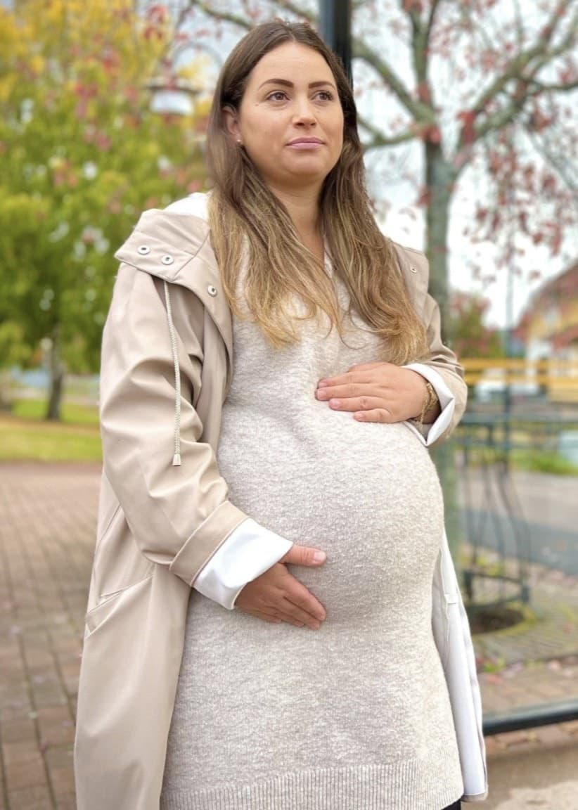 En kvinna runt 30 år som är höggravid och som står vid en busshållplats och håller händerna på sin mage.