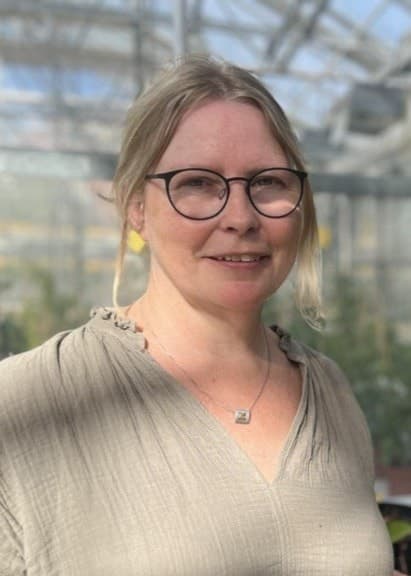 Kvinna står i ett växthus omgiven av gröna plantor i krukor. Solljus tränger igenom glasväggarna och skapar en ljus och naturlig miljö.