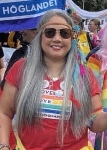 Sofia Rudmark med regnbågsflaggor deltar i en Pride-parad. I bakgrunden syns fler deltagare och färgglada flaggor. En prideflagga med texten ”Love is love Höglandet” är synlig.