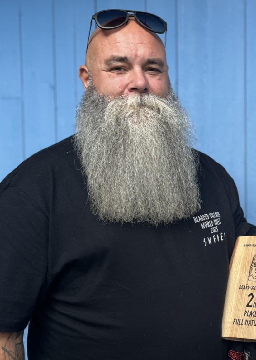 Daniel Bergström med långt grått skägg står framför en blå vägg, klädd i svart t-shirt och håller en träskylt med texten 'Best Beard'.