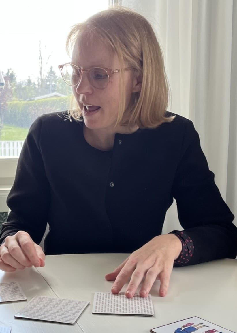 Linda Johansson sitter vid ett bord med två barn och spelar ett sällskapsspel. Bordet är täckt med kort och papper, och ett fönster med utsikt över villor syns i bakgrunden.