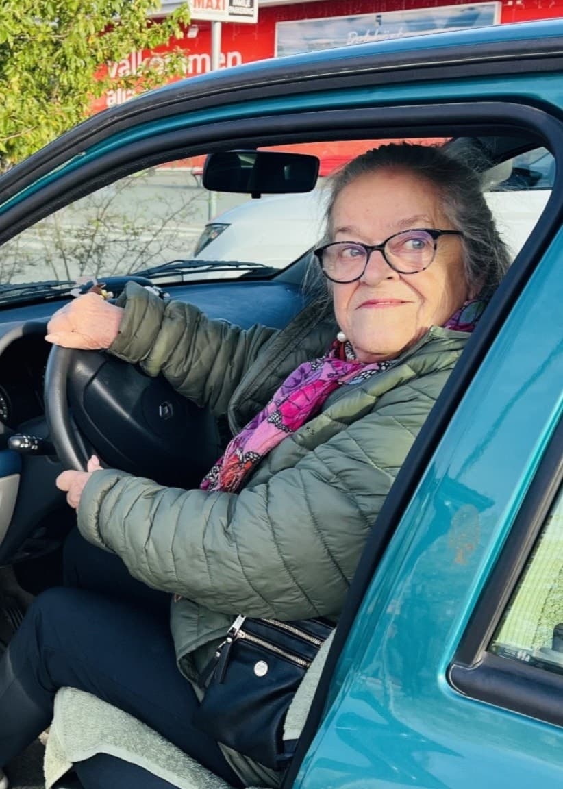 En person sitter i förarsätet på en grön bil som står parkerad utomhus vid en så kallad familjeparkering utanför en större butik. Bakgrunden visar träd och en byggnad med röda detaljer.