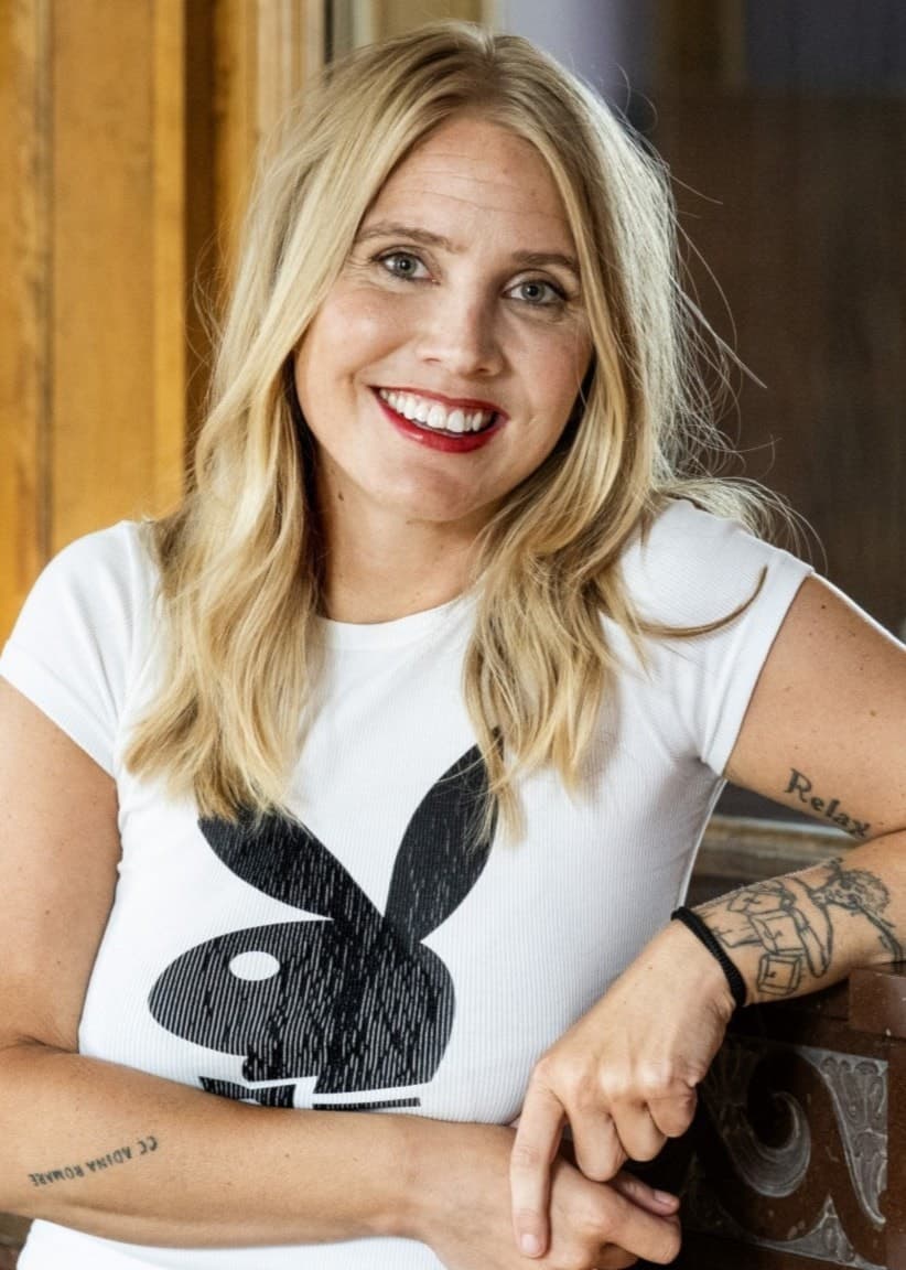 Författaren Amanda Romare står lutad mot en träpanel i en inomhusmiljö. Hon har långt blont hår, en vit t-shirt med ett svart playboy-bunny-tryck och en tatuering på ena armen.