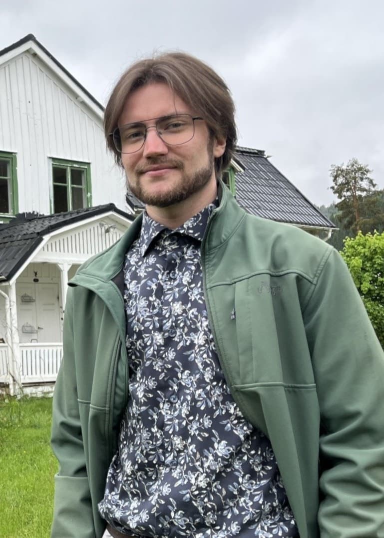 En man står framför ett vitmålat hus med gröna fönsterkarmar och en mindre klocktornsliknande byggnad på taket, omgiven av grönskande trädgård under en molnig himmel.