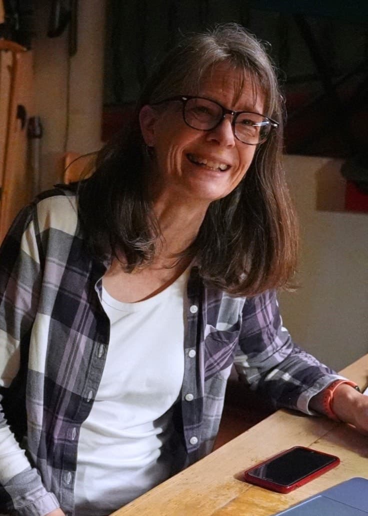Mary E. Brunkow, klädd i en rutiga skjorta, sitter vid ett köksbord med en laptop, mobil och kaffemugg, och ler efter att ha fått veta att hon vunnit Nobelpriset i medicin för forskning inom immunologi.