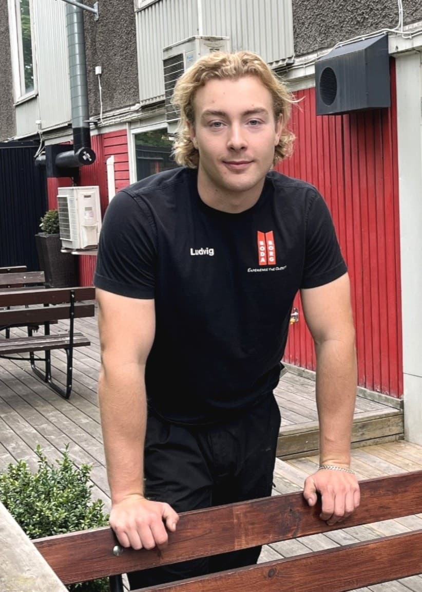 Ludvig Persson, personal på Boda Borg, står utomhus vid en träbänk framför en byggnad med stora glasfönster och röda väggar. Gräs och buskar syns i bakgrunden.