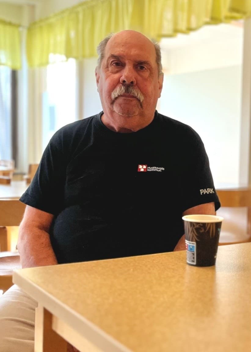 Mats Wedin sitter vid ett träbord i en matsal med en papperspåse, kaffemugg och en mobiltelefon framför sig. Ljusa gardiner och trästolar syns i bakgrunden.
